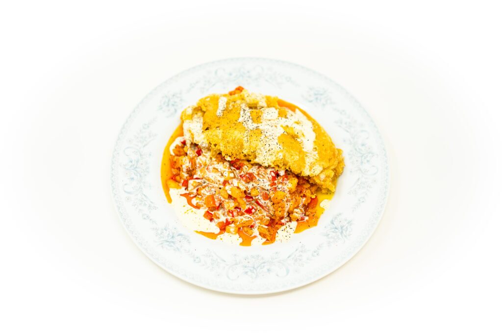 A plate of crispy masa beer-battered grouper resting on a bed of diced chayote and potato hash, finished with a heavy pour of white lime crema and bright red chili oil.