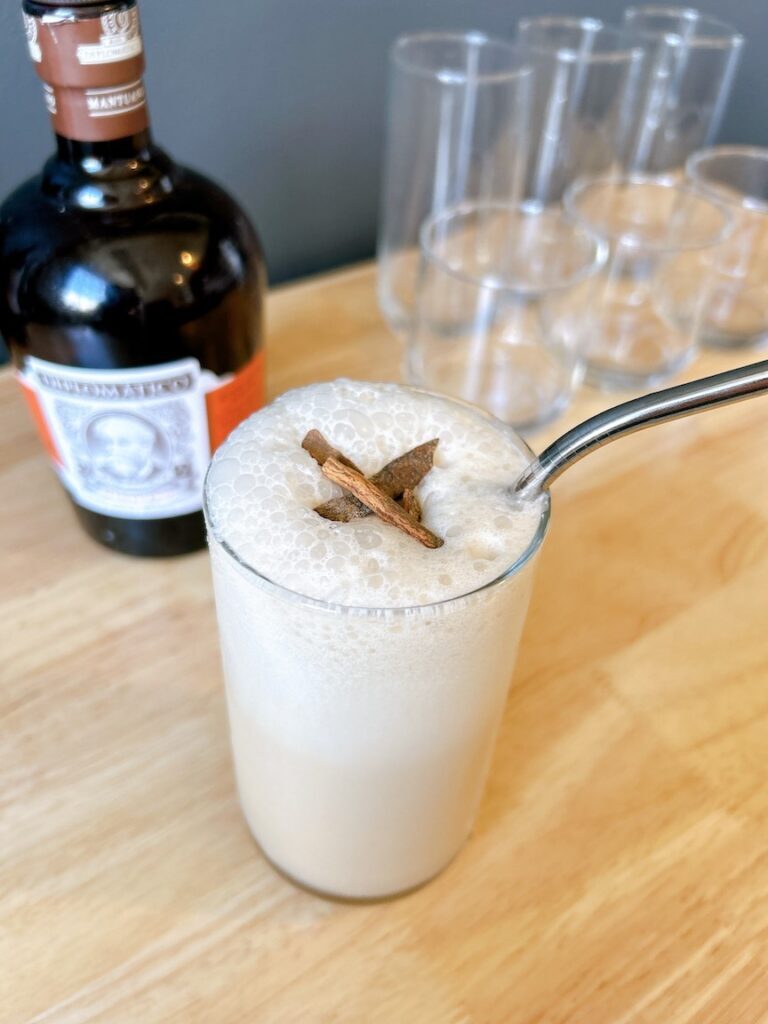 Iced coconut milk rum latte served in a clear glass with a cinnamon stick, on a wooden table beside a bottle and empty glasses.