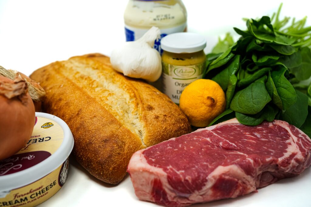 Ingredients for a New York steak sandwich including a raw steak, ciabatta bread, spinach, red onion, a lemon, and jars of seasoning arranged on a white background.