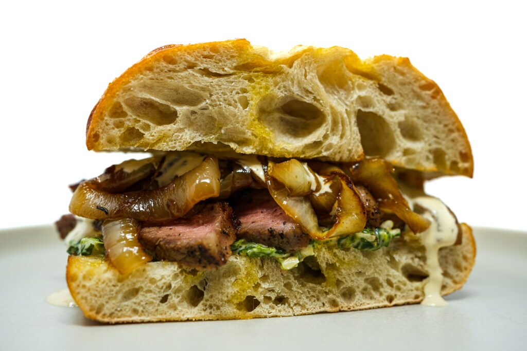 A ny strip steak sandwich with tender slices of seared steak on a toasted bun, topped with creamy spinach and caramelized onions, drizzled with steak sauce.