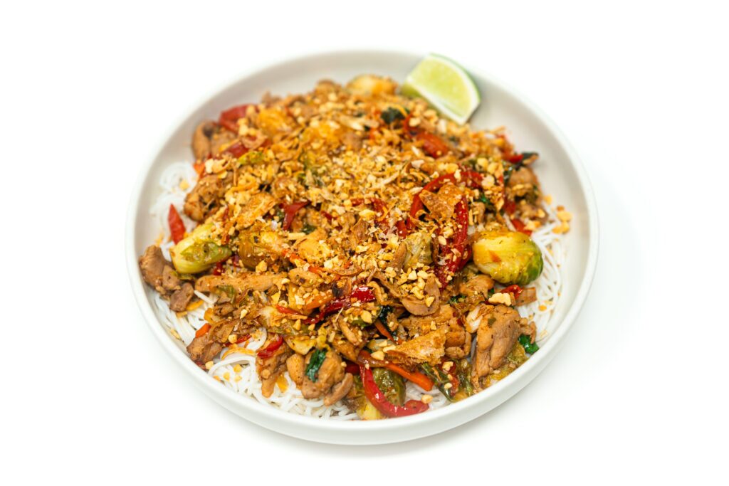 A vibrant plate of Thai Chili Chicken & Brussels Sprouts Stir-Fry, featuring golden seared chicken, crispy Brussels sprouts, red bell peppers, and Thai chilies, all coated in a glossy, umami-rich sauce and garnished with fresh basil, cilantro, and crushed peanuts.