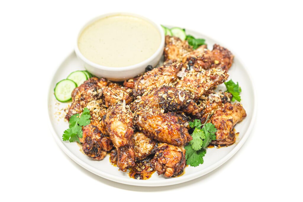 A plate of crispy Hot Honey Garlic Wings, coated in a sticky, spicy-sweet glaze, served with a side of creamy Cilantro Lime Ranch and garnished with fresh cilantro and lime wedges.