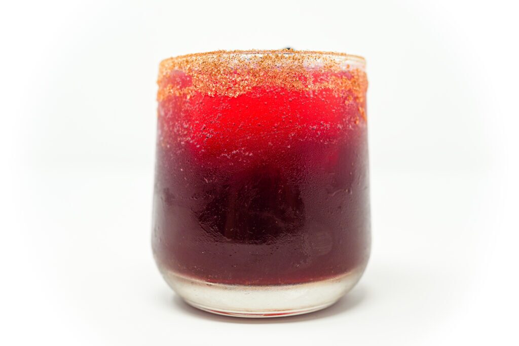 A cinnamon hibiscus mezcal margarita in a glass rimmed with cinnamon, showcasing a deep red hue from the hibiscus infusion. The drink is elegantly garnished with a cinnamon stick and a vibrant red hibiscus flower, enhancing its visual appeal.