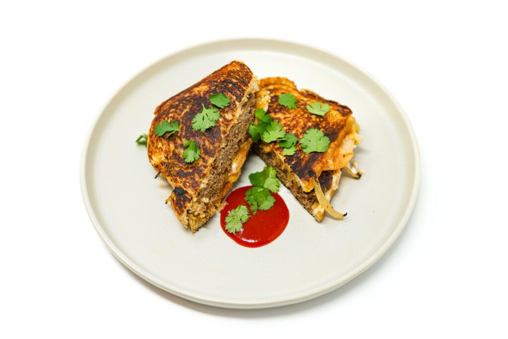 A Korean BBQ Patty Melt on a plate, showcasing a juicy beef patty with caramelized onions and kimchi, melted cheese between toasted sourdough slices, served with a side of sriracha sauce for dipping.