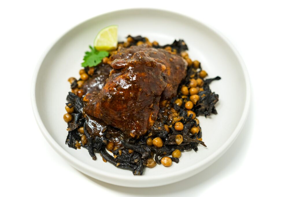 A plate of Chicken Mole with Sautéed Peas and Mushrooms featuring tender chicken thighs in a rich mole sauce, served over a bed of peas and sautéed mushrooms with shallots and garlic, garnished with lime wedges.