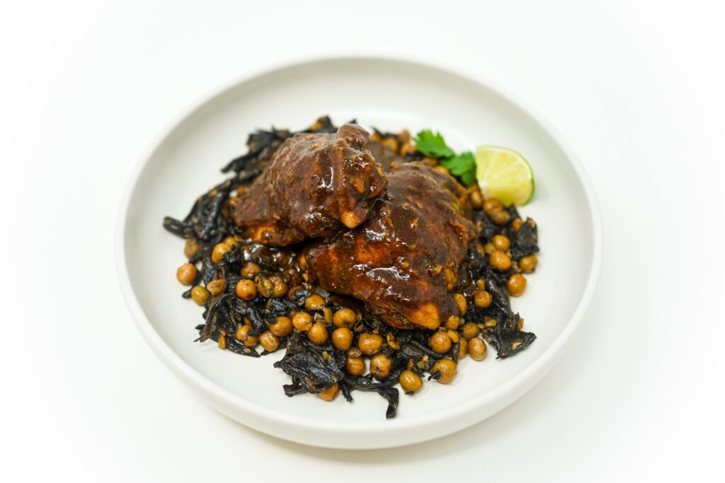 A plate of Chicken Mole with Sautéed Peas and Mushrooms featuring tender chicken thighs in a rich mole sauce, served over a bed of peas and sautéed mushrooms with shallots and garlic, garnished with lime wedges.
