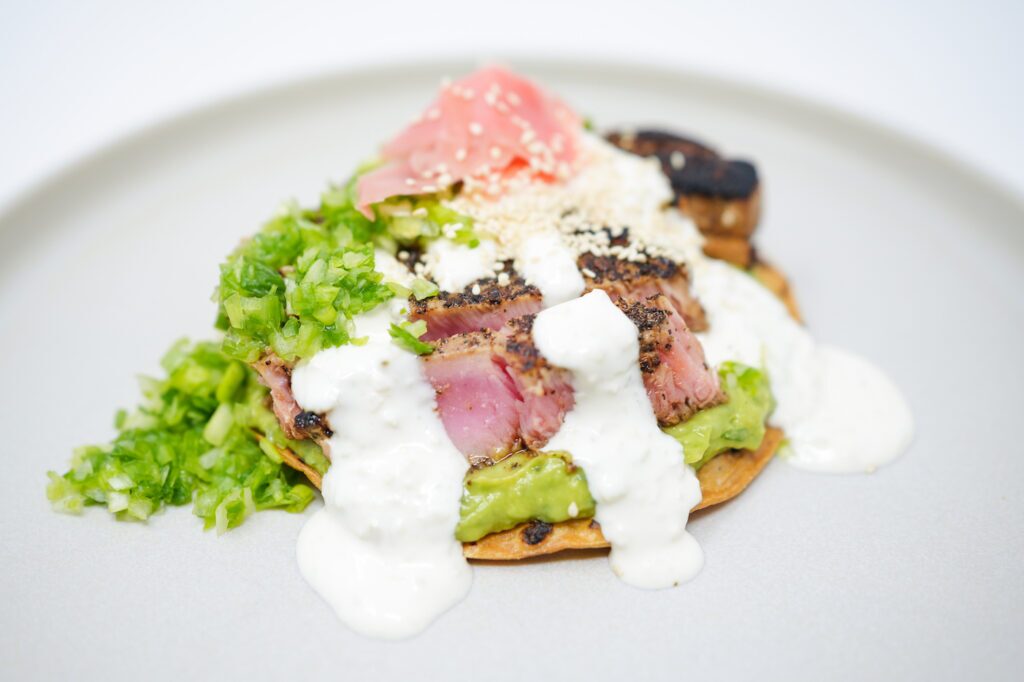 A Seared Ahi Tuna Tostada with a crispy tortilla topped with marinated seared ahi tuna slices, creamy wasabi guacamole, drizzles of garlic soy aioli, garnished with chopped green onions, toasted sesame seeds, and pickled ginger.