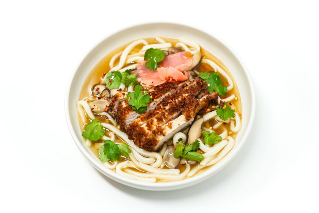 A bowl of Don Katsu Pork Udon Noodle Soup featuring golden, crispy pork cutlets atop a bed of thick udon noodles in a rich, smooth ginger broth, garnished with fresh cilantro and pickled ginger