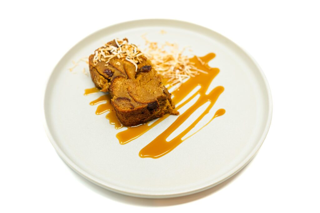 A slice of Coconut Caramel Raisin Bread Pudding drizzled with rich caramel sauce and garnished with toasted coconut flakes, showcasing its golden-brown crust and creamy interior