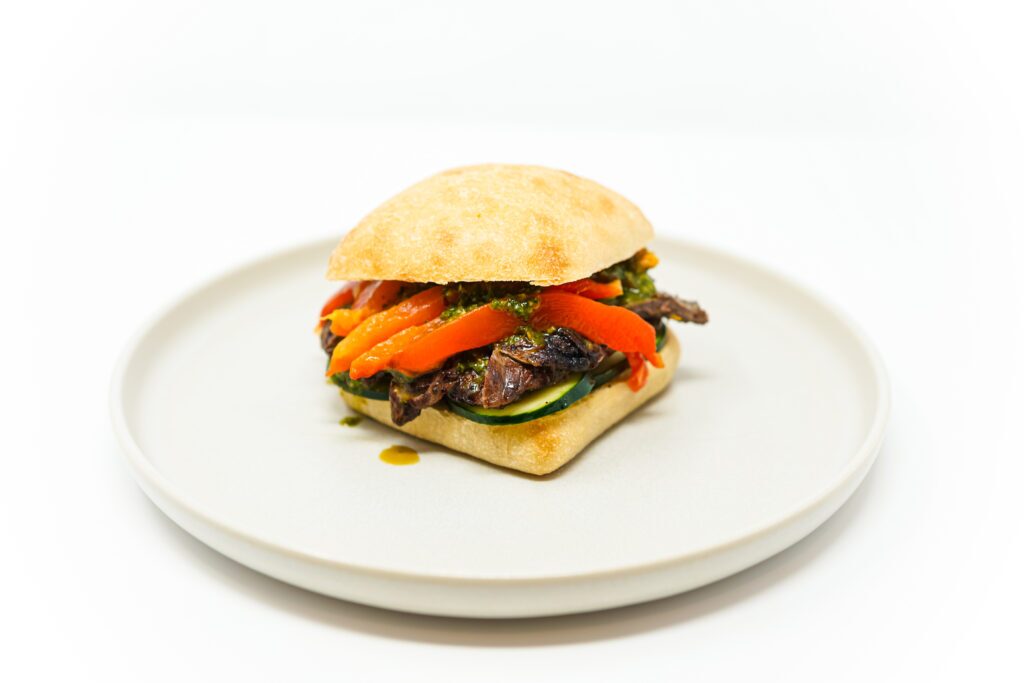 A close-up image of a Grilled Flank Steak Chimichurri Sandwich featuring juicy slices of marinated flank steak, vibrant green chimichurri sauce, roasted red pepper strips, and fresh cucumber slices layered on a toasted ciabatta roll.