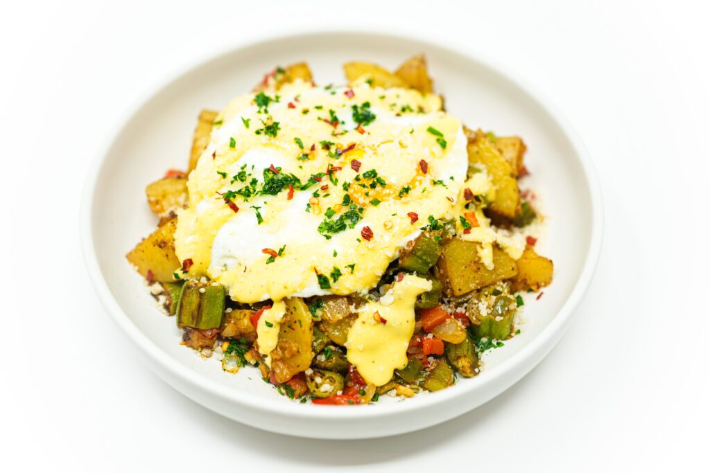 Southern okra hash with crispy potatoes, sautéed vegetables, a fried egg, and creamy tahini aioli, garnished with parsley and chili flakes.