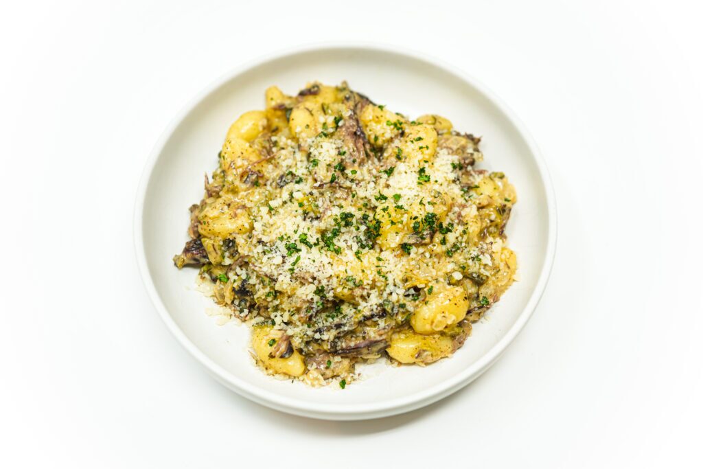 A plate of creamy gnocchi topped with shredded smoky brisket and roasted poblano peppers, garnished with fresh cilantro and a sprinkle of Parmesan cheese. The dish has a rich, golden sauce and is served with a dash of paprika for added color.