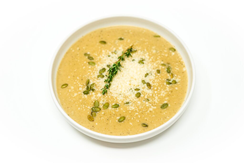 A bowl of creamy roasted garlic and white pumpkin bisque, garnished with toasted pumpkin seeds, fresh thyme, and grated Parmesan cheese.
