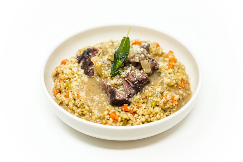 A plate of Sage-Braised Short Ribs served over pearl barley, topped with a rich, sage-infused pan gravy and garnished with fresh herbs for a rustic and comforting presentation.