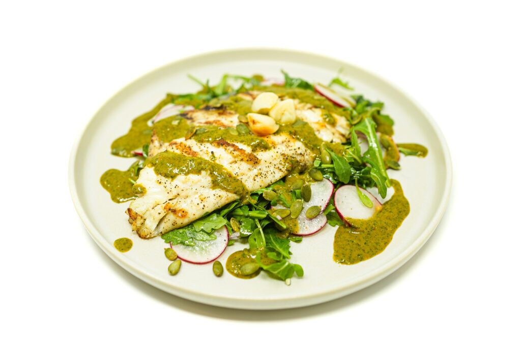 Grilled speckled trout with roasted garlic chimichurri, served on a bed of arugula, radishes, and toasted pumpkin seeds on a neutral plate.