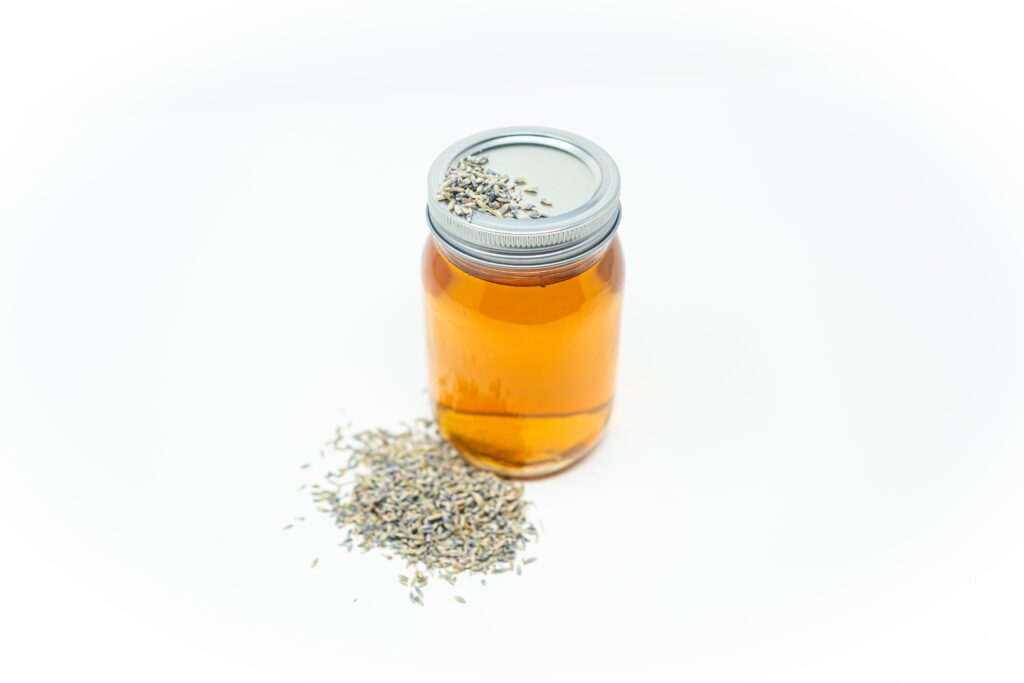 A glass jar filled with golden lavender-infused vodka, placed on a white background with dried lavender buds scattered beside it.