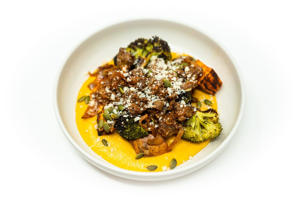 A shallow bowl of French-style ground lamb served over brown butter butternut squash purée with roasted broccoli and sweet potatoes, topped with Parmesan and herbs.
