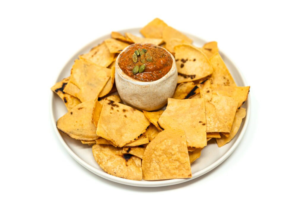 A plate of homemade tortilla chips surrounding a small bowl filled with roasted tomato and red pepper salsa garnished with pumpkin seeds.