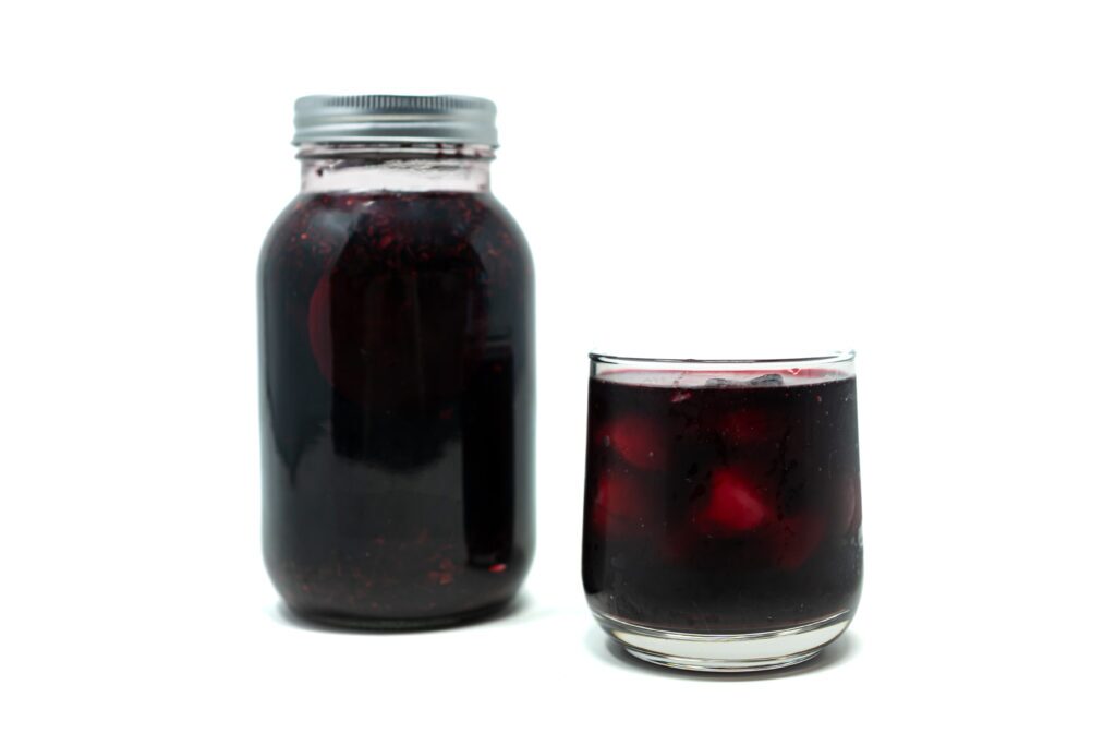 A glass jar of ruby-red hibiscus tea with lemon slices and ginger on a white surface, photographed in natural light.