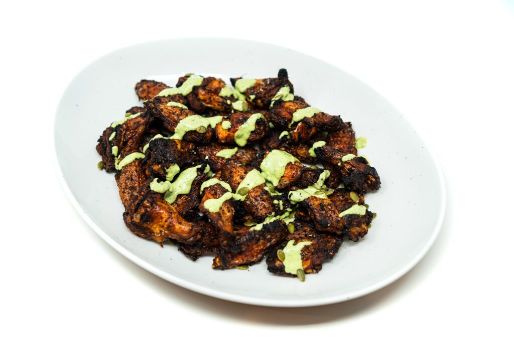 Close-up of jerk-rubbed chicken wings with creamy jalapeño herb sauce, garnished with herbs and lime zest on a rustic plate.