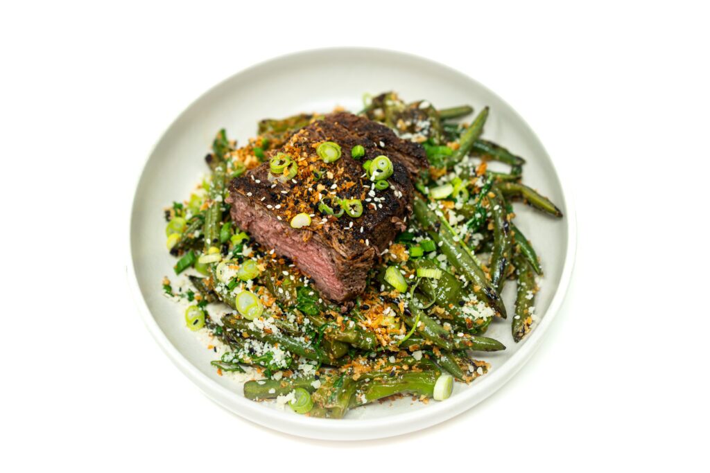 Grilled and sliced New York strip steak layered over a Caesar salad with charred shishito peppers, green beans, arugula, and toasted panko.