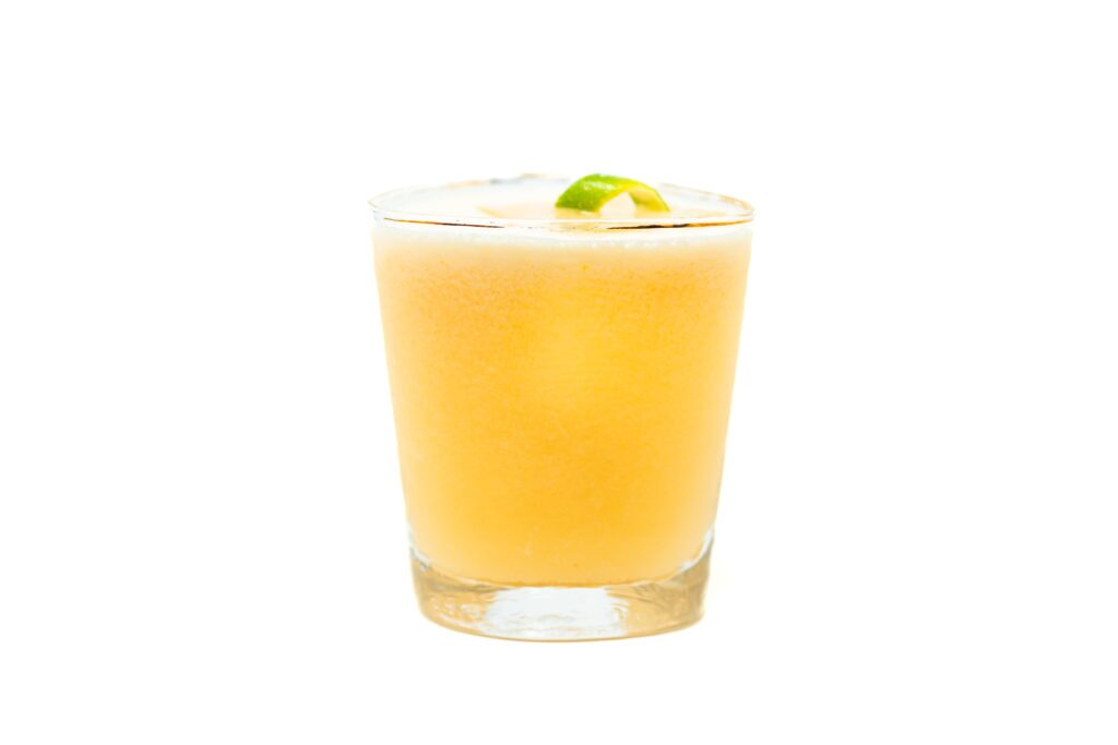 A golden orange persimmon milk punch margarita served in a rocks glass with a lime wedge garnish, showing a silky and slightly frothy top.