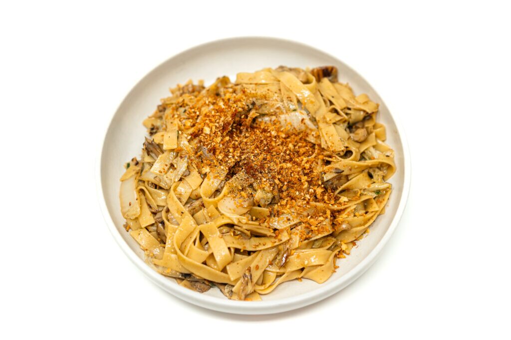 A white bowl containing wok-charred maitake mushroom and napa cabbage tagliatelle pasta tossed in a soy butter sauce and topped with golden toasted sesame panko breadcrumbs.