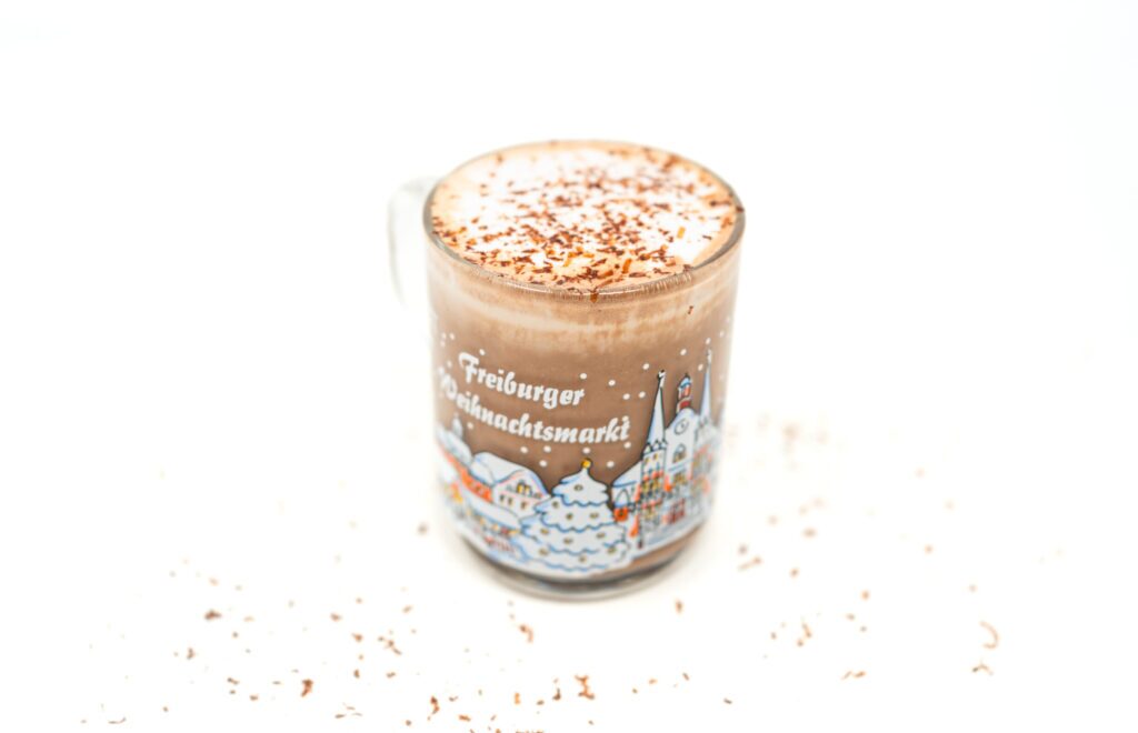 A clear glass mug filled with rich hot chocolate latte sits on a white surface surrounded by chocolate shavings, featuring a layer of frothy cream on top and a colorful illustration of a snowy town on the glass.