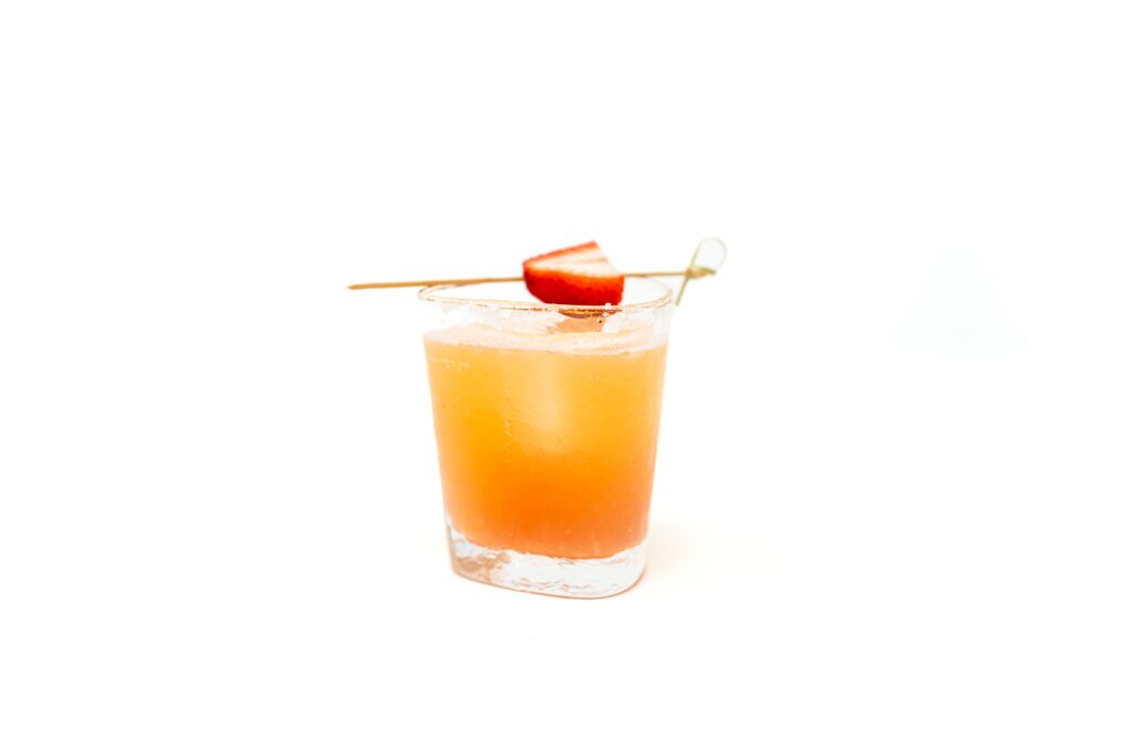 An overhead view of a strawberry margarita in a modern glass with a large clear ice cube, garnished with a fresh strawberry slice on a wooden skewer