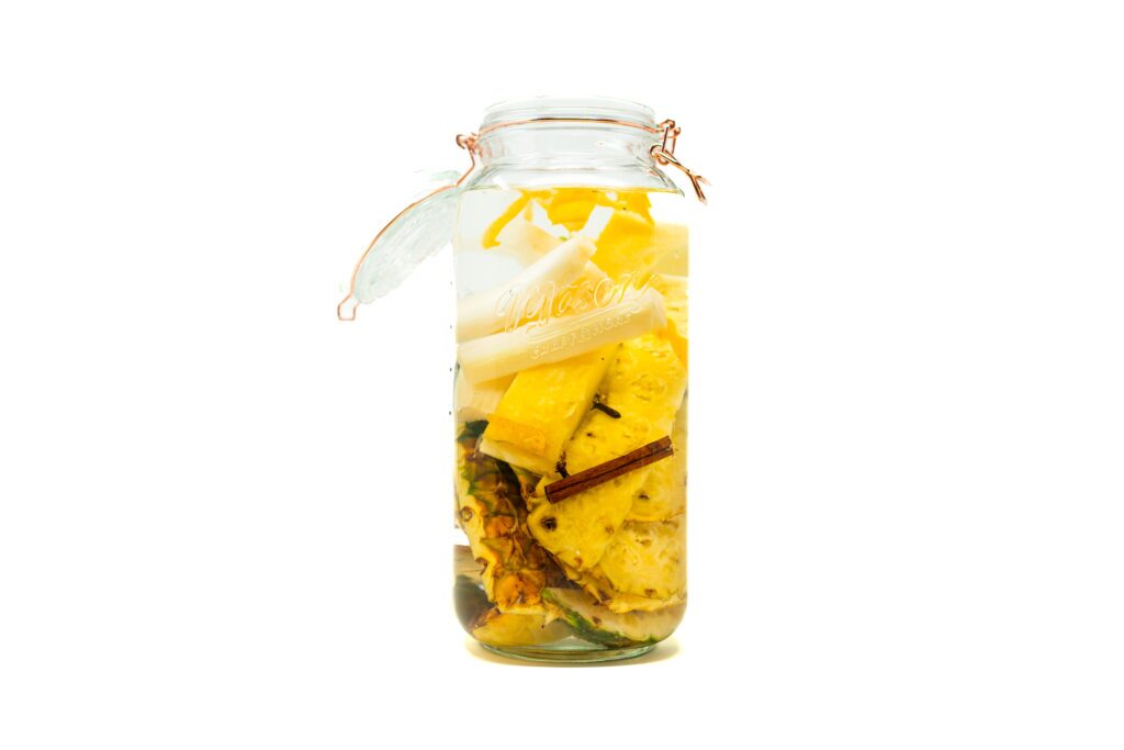 A glass jar filled with pineapple rinds, fresh sugar cane batons, and cinnamon sticks submerged in water for fermentation.