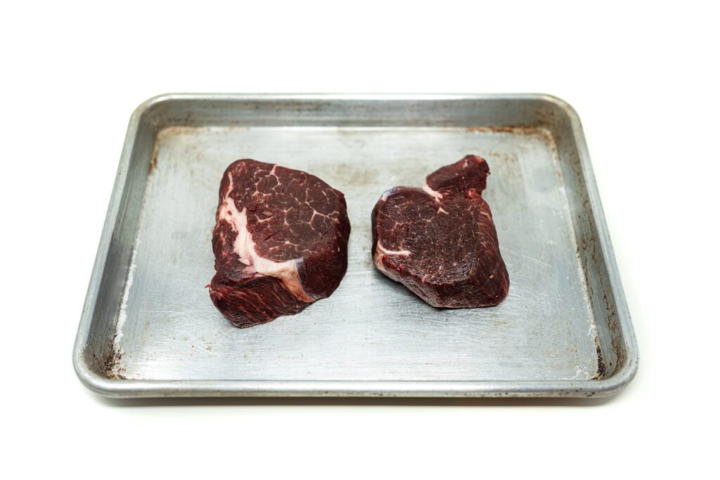 Two dry-brined filet mignon steaks resting on a metal sheet pan against a white background.