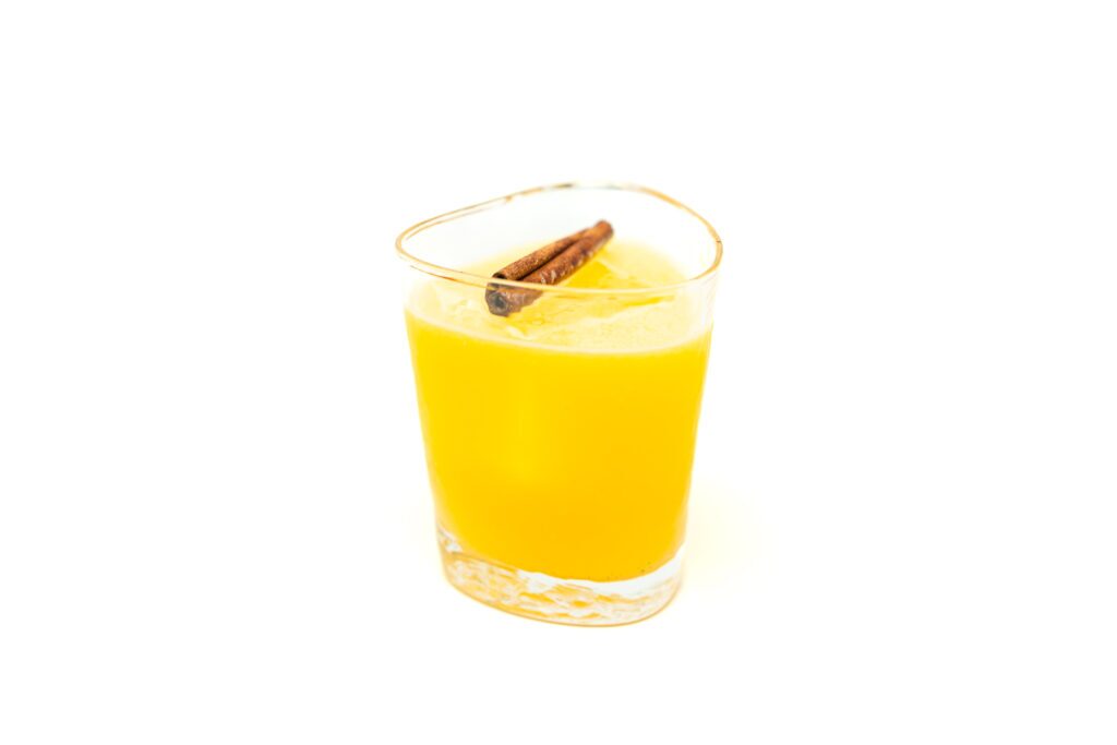 A rocks glass filled with a bright orange Mezcal Coffee Orange Cocktail garnished with a bruised cinnamon stick resting on fresh ice.