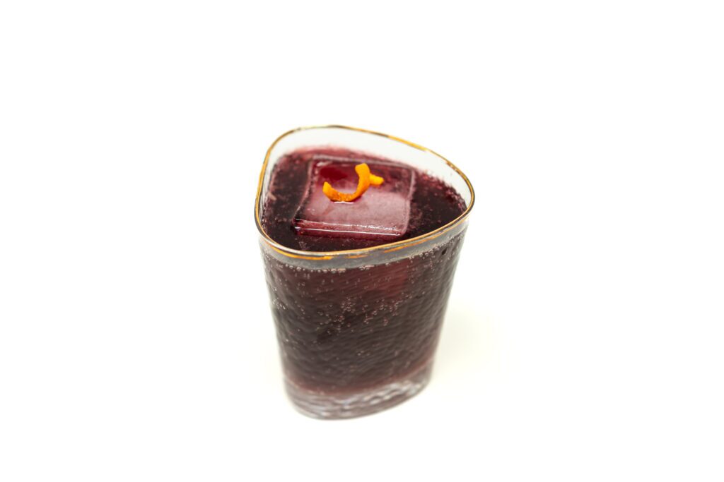 A dark red wine spritzer with a hazy orange glow, garnished with an orange twist and served next to a bottle of Halozan red wine.
