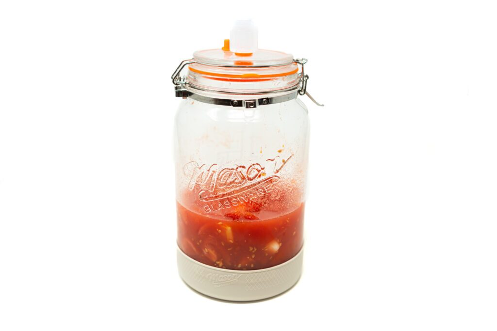 A glass fermentation jar holding dry-macerated tomatoes, garlic, and vines submerged in their natural liquid under an airlock lid.