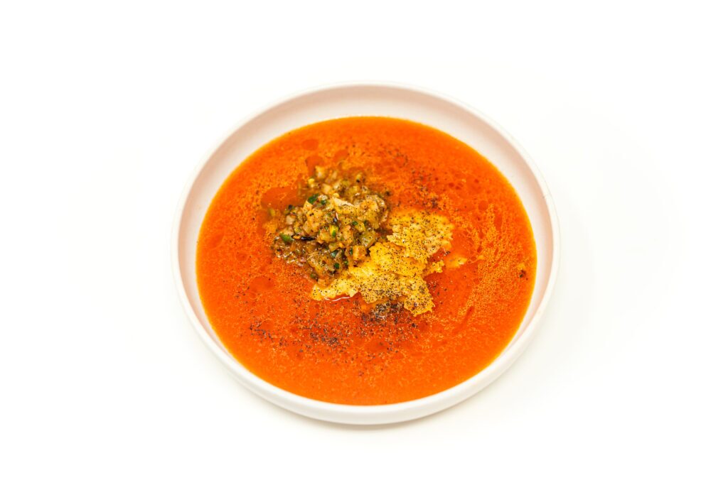 A shallow bowl of bright orange lacto-fermented gazpacho topped with a mound of wok-charred fennel relish and a jagged shard of crispy parmesan.