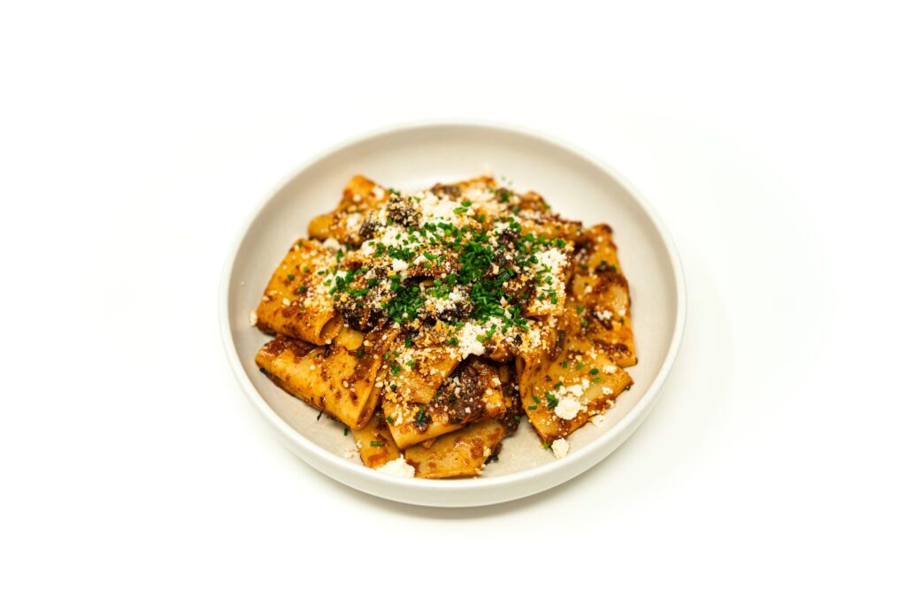 A wide bowl filled with paccheri pasta coated in a dark braised oxtail ragu, finished with grated cheese and razor-thin chives.