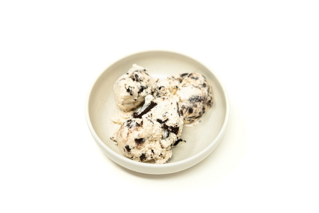 A close up shot of a scoop of dense, speckled cookies and cream ice cream with large chunks of chocolate sandwich cookies in a dark ceramic bowl.