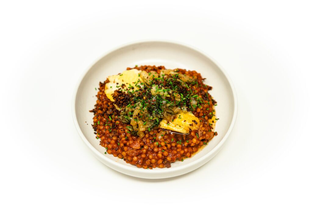 A perfectly plated pan seared haddock fillet resting over rich fire-roasted tomato pearl couscous, topped with charred bok choy salad and a golden ginger allium crunch in a white ceramic bowl