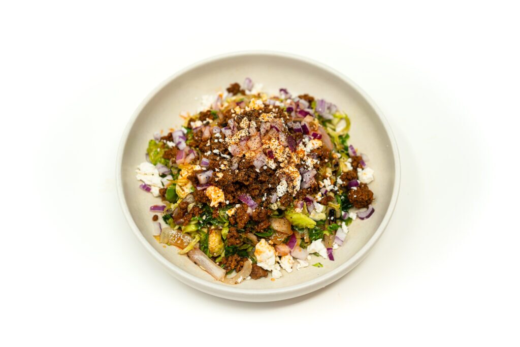 A shallow bowl of spiced cumin lamb salad resting on a bed of charred endive and Brussels sprouts, finished with red onion and crumbled feta.