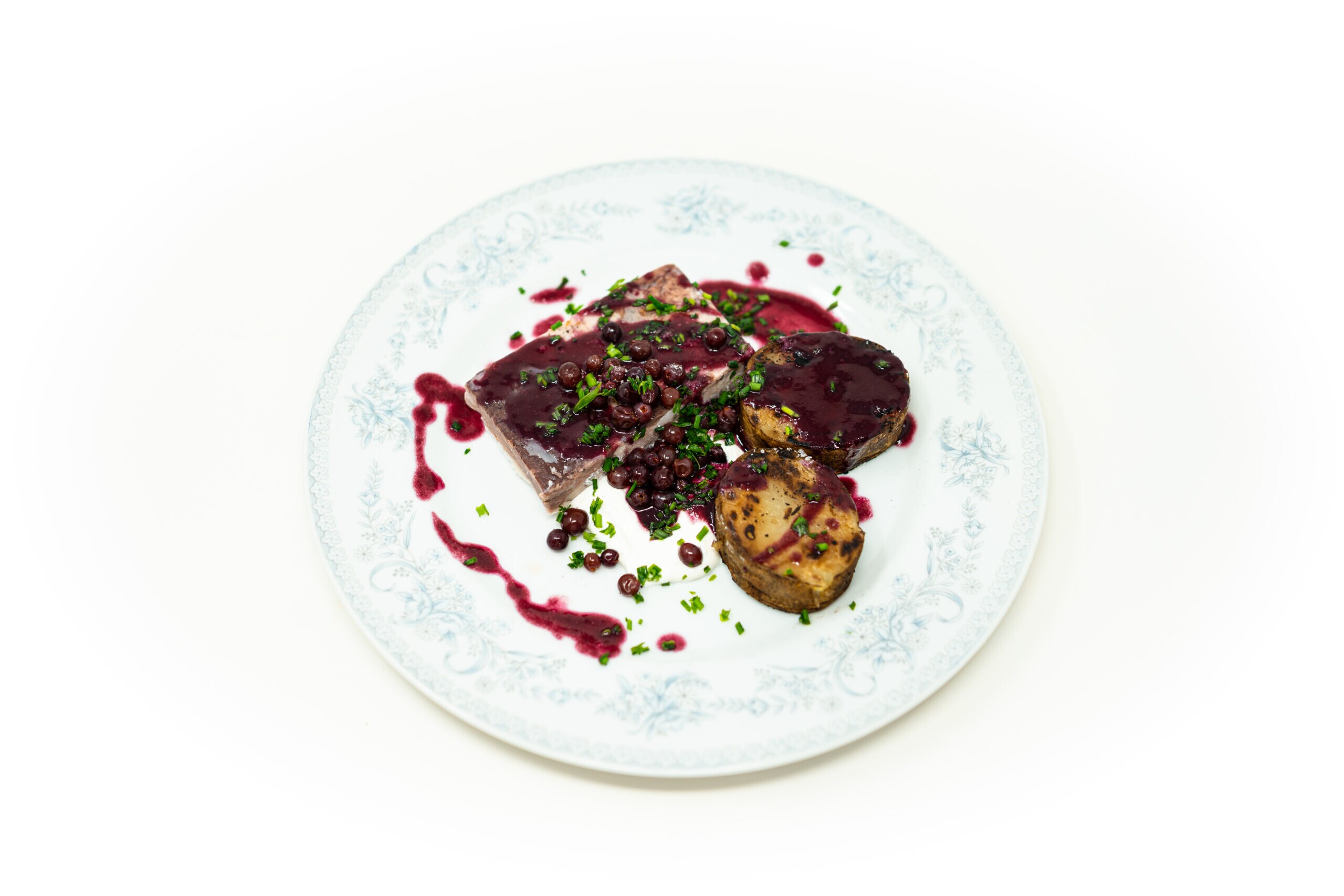 Top-down view of sliced compressed pork belly glossed in dark purple Concord agrodolce alongside charred thick-cut potatoes and bright green chives.