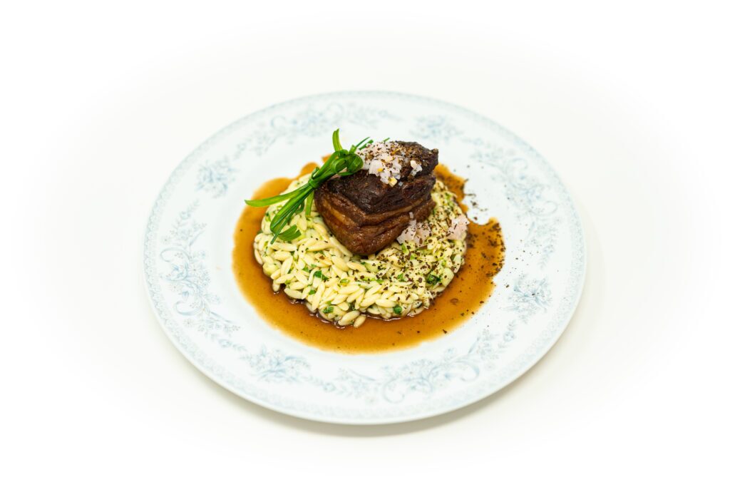 Overhead view of square-cut kombu glazed pork belly topped with pickled shallots and tied chives on a bed of herb-flecked sushi-spiced orzo surrounded by dark reduction sauce on a patterned white plate.