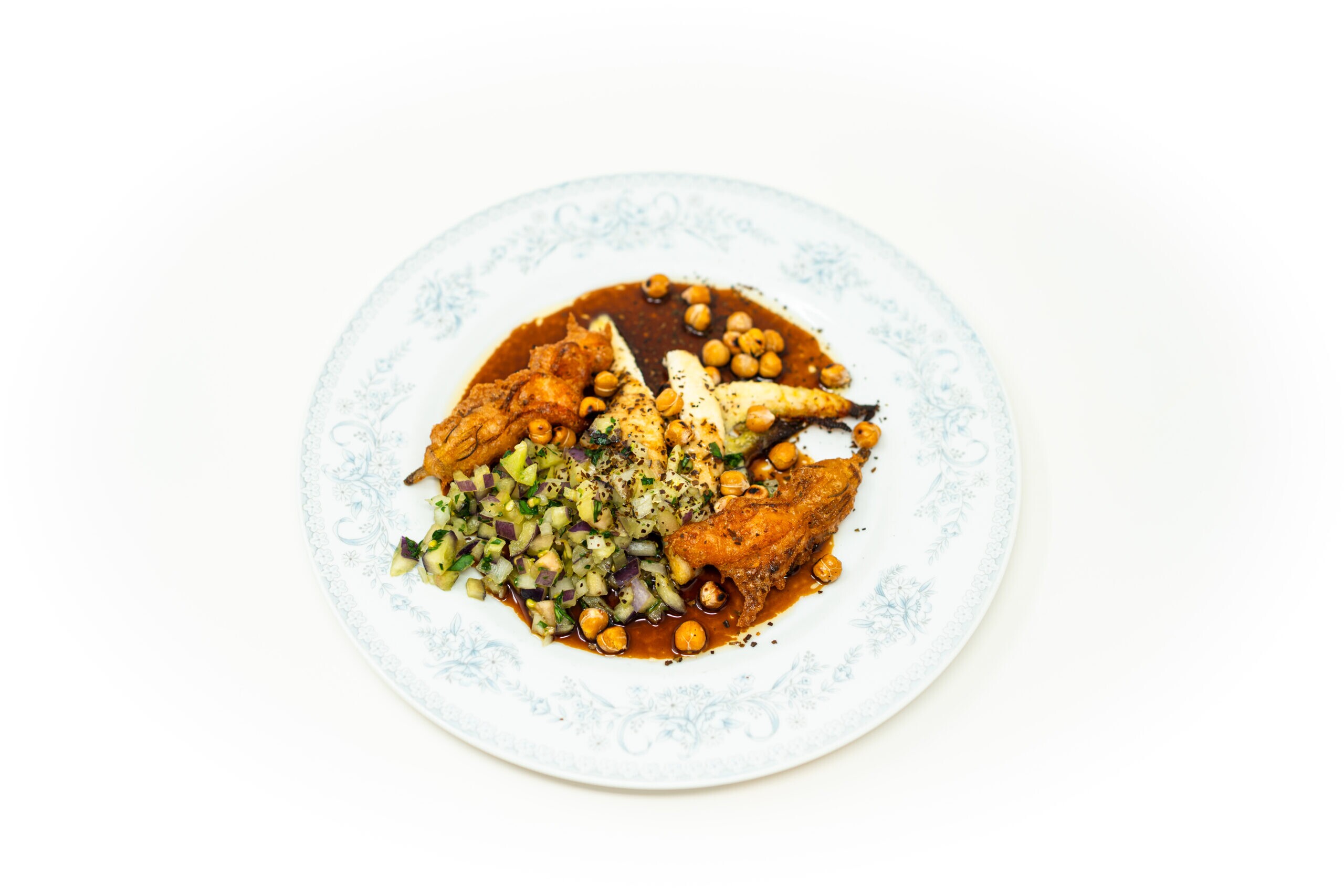 Close-up angled shot of pan-fried flatfish split to reveal its white steamed interior, surrounded by dark brown agrodolce sauce, raw purple pepper brunoise, fried squash blossoms, and blistered chickpeas under bright, hard lighting.