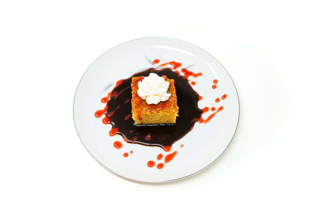 A square slice of golden semolina cake rests in a pool of dark muscovado caramel dotted with red strawberry puree, topped with a white cream quenelle.