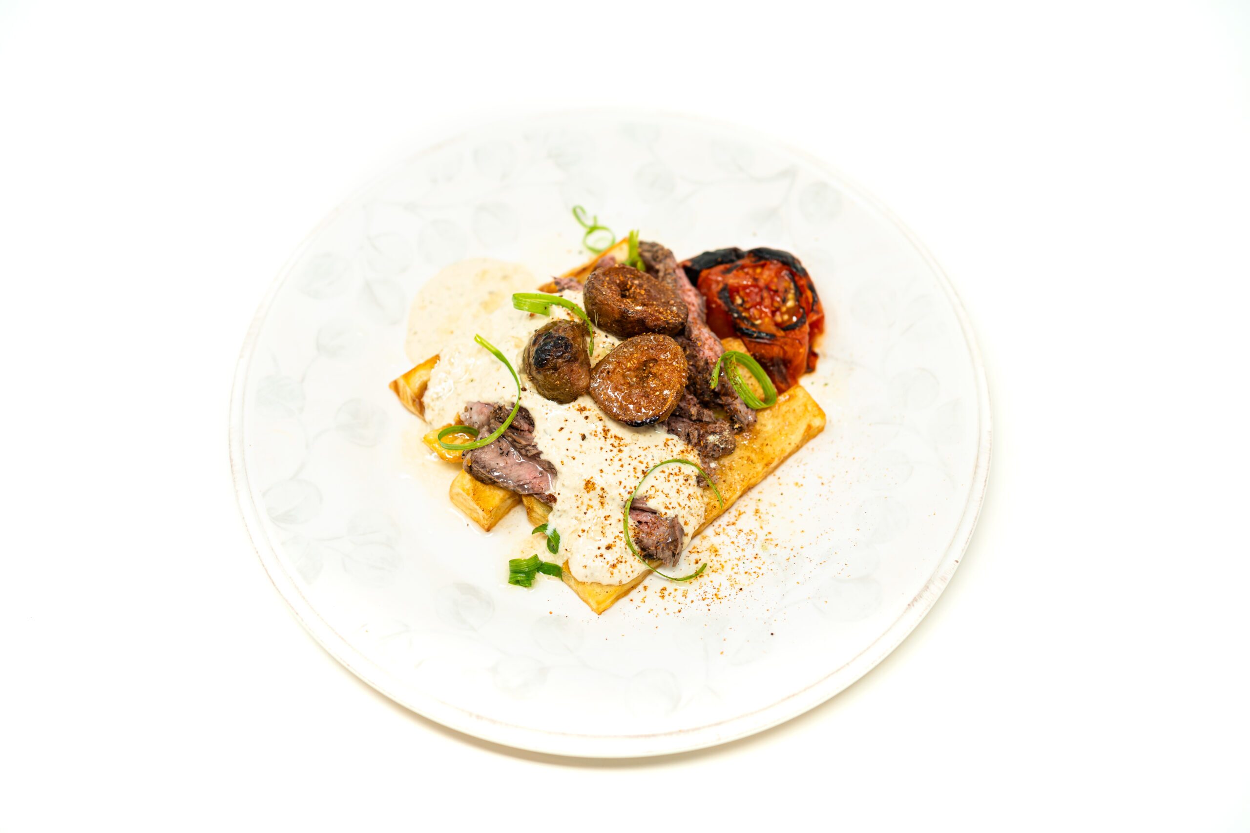 Overhead plated view of a seared ribeye resting on crispy blanched potato wedges, covered in green peppercorn tartare relish and tomato skin powder.